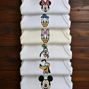 May include: A stack of white baby onesies featuring embroidered Disney character faces. The characters include Minnie Mouse, Donald Duck, Daisy Duck, Goofy, Pluto, and Mickey Mouse. The onesies are stacked on a dark wood surface.