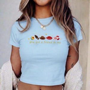 May include: A light blue short-sleeved t-shirt featuring a graphic print of a colorful ball, a boot, a brown cowboy hat, a red hat, and a rocket ship. The text "You got a friend in me" is printed in yellow below the icons with small yellow stars.