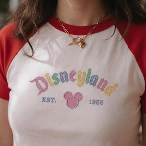 Op de afbeelding: Wit en rood raglan T-shirt met het woord "Disneyland" geborduurd in pastelkleuren. Het shirt heeft een Mickey Mouse-hoofd en de tekst "EST. 1955". Een gouden ketting met een hartvormige hanger wordt gedragen.