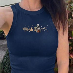 Puede incluir: Camiseta sin mangas acanalada azul marino con cuello redondo. La camiseta presenta un peque&ntilde;o gr&aacute;fico de personajes y s&iacute;mbolos de una popular franquicia de ciencia ficci&oacute;n. El gr&aacute;fico incluye una nave espacial, un s&iacute;mbolo, un personaje que sostiene a un beb&eacute; y un caza estelar.