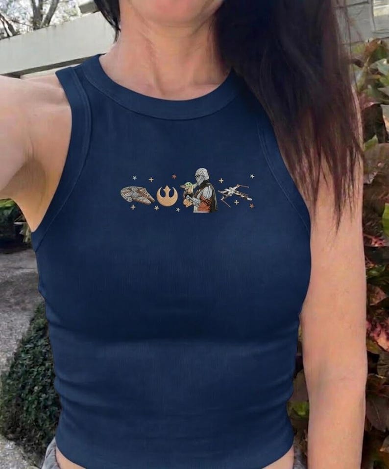 Puede incluir: Camiseta sin mangas acanalada azul marino con cuello redondo. La camiseta presenta un peque&ntilde;o gr&aacute;fico de personajes y s&iacute;mbolos de una popular franquicia de ciencia ficci&oacute;n. El gr&aacute;fico incluye una nave espacial, un s&iacute;mbolo, un personaje que sostiene a un beb&eacute; y un caza estelar.