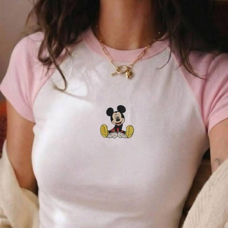 May include: White and pink raglan style t-shirt featuring a small embroidered image of Mickey Mouse. The shirt has a white body with pink sleeves and neckline. The cartoon character is sitting with his legs crossed.