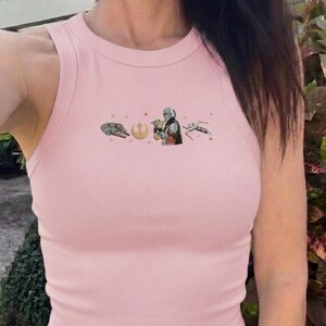 Puede incluir: Una camiseta sin mangas rosa claro con costuras acanaladas y un gr&aacute;fico del Halc&oacute;n Milenario, un s&iacute;mbolo, El Mandaloriano sosteniendo a Grogu y un caza X-wing. La camiseta tiene cuello redondo y corte ajustado.