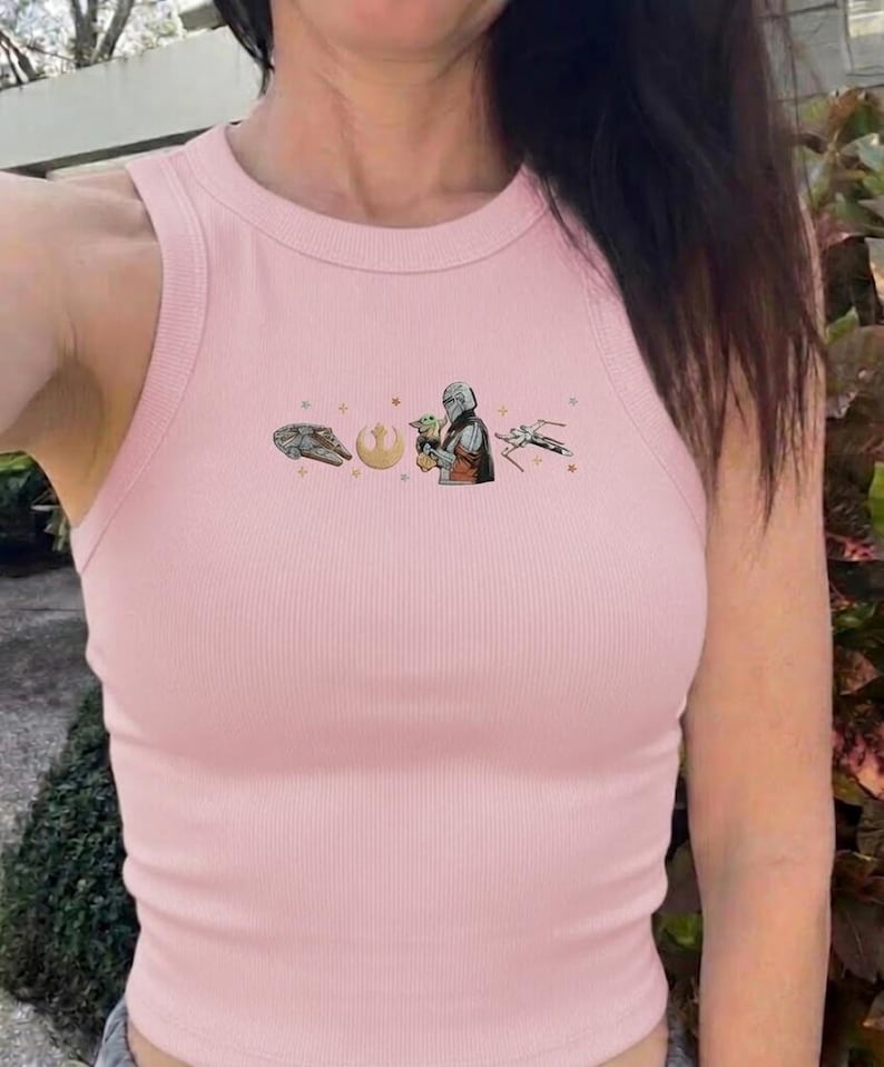Puede incluir: Una camiseta sin mangas rosa claro con costuras acanaladas y un gr&aacute;fico del Halc&oacute;n Milenario, un s&iacute;mbolo, El Mandaloriano sosteniendo a Grogu y un caza X-wing. La camiseta tiene cuello redondo y corte ajustado.