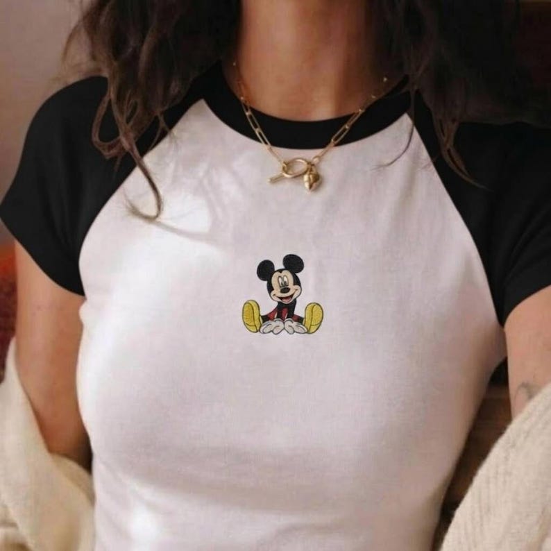 May include: White raglan t-shirt with black sleeves featuring a seated Mickey Mouse graphic. The cartoon character is depicted in his classic red shorts, yellow shoes, and black ears. The shirt is a casual style.