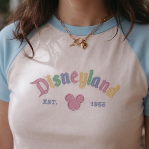 Op de afbeelding: Wit en lichtblauw raglan T-shirt met het woord "Disneyland" geborduurd in pastelkleuren. Het shirt heeft ook een roze Mickey Mouse-hoofd en de tekst "EST. 1955". Een gouden ketting met een sleutel- en hartvormige hanger wordt gedragen.