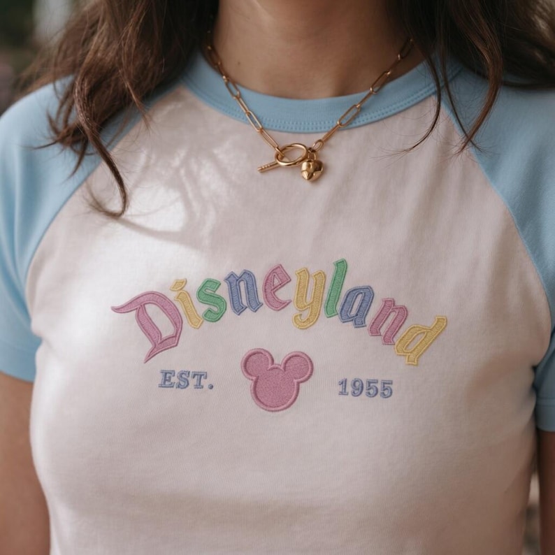 Op de afbeelding: Wit en lichtblauw raglan T-shirt met het woord "Disneyland" geborduurd in pastelkleuren. Het shirt heeft ook een roze Mickey Mouse-hoofd en de tekst "EST. 1955". Een gouden ketting met een sleutel- en hartvormige hanger wordt gedragen.