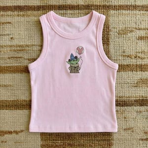 Puede incluir: Una camiseta sin mangas rosa claro con cuello redondo y un dibujo animado de un personaje verde con un sombrero de mago y un globo de Mickey Mouse. La camiseta sin mangas est&aacute; hecha de un material suave y acanalado.