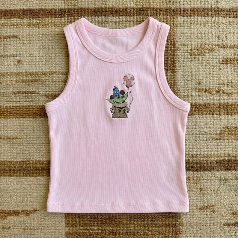 Puede incluir: Una camiseta sin mangas rosa claro con cuello redondo y un dibujo animado de un personaje verde con un sombrero de mago y un globo de Mickey Mouse. La camiseta sin mangas est&aacute; hecha de un material suave y acanalado.