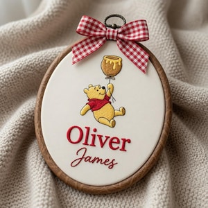 Könnte beinhalten: Ein ovaler Stickrahmen mit weißem Stoffhintergrund zeigt einen gestickten gelben Bären im roten Hemd, der einen braunen Honigtopf-Ballon hält. Unter dem Bären ist der Name Oliver James in Rot gestickt. Eine rot-weiß karierte Schleife ist oben befestigt.