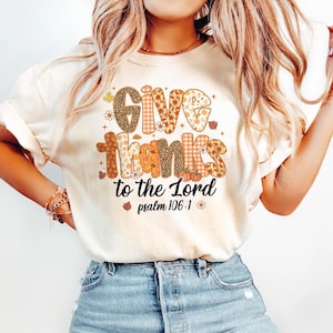以下が含まれることがあります： 秋をテーマにしたデザインで「Give Thanks to the Lord」という言葉がプリントされたクリーム色のTシャツ。テキストには、花柄、ヒョウ柄、チェック柄が含まれています。「詩篇106：1」というフレーズもシャツにプリントされています。