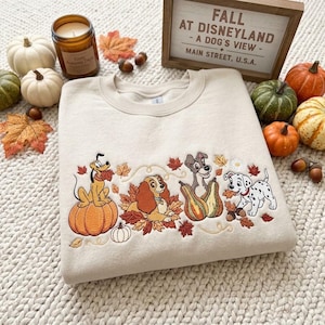 May include: A beige crewneck sweatshirt featuring an embroidered design of four Disney dogs—Pluto, Lady, Tramp, and a Dalmatian puppy—surrounded by autumn leaves and pumpkins. A wooden sign above reads FALL AT DISNEYLAND - A DOG'S VIEW - MAIN STREET, U.S.A.