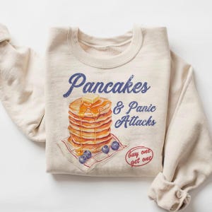 May include: Beige sweatshirt with the text "Pancakes & Panic Attacks" in blue script. The graphic features a stack of pancakes with butter and syrup, blueberries, and a "buy one get one" stamp.
