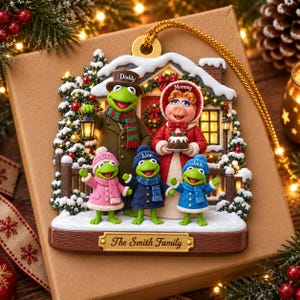 May include: A decorative Christmas ornament featuring a frog family in front of a house. The figures are labeled Daddy, Mommy, Lisa, Lion, and Jack. The base displays the text The Smith Family. The scene includes snow, trees, and a lantern.