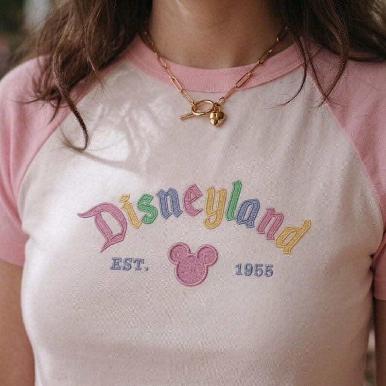 Op de afbeelding: Een wit en roze raglan T-shirt met het woord "Disneyland" geborduurd in pastelkleuren. Het shirt heeft een Mickey Mouse-hoofd en de tekst "EST. 1955". Een gouden ketting met een hartvormige hanger wordt gedragen.
