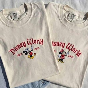 May include: Two off-white t-shirts with the embroidered text "Disney World" in red script, along with the year "1971". One shirt features Mickey Mouse, the other Minnie Mouse, both holding paintbrushes. The shirts are laid on a white surface.