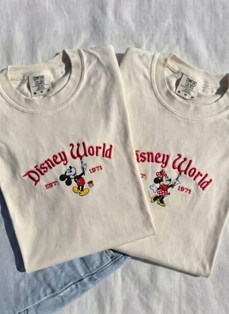 May include: Two off-white t-shirts with the embroidered text "Disney World" in red script, along with the year "1971". One shirt features Mickey Mouse, the other Minnie Mouse, both holding paintbrushes. The shirts are laid on a white surface.