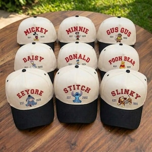 May include: A collection of baseball caps with white crowns and black brims. Each cap features a different Disney character's name embroidered in red, along with the character's image and the year they were established.