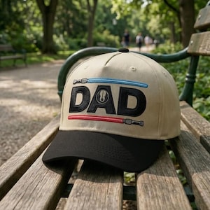 May include: A tan baseball cap with a black brim featuring the word "DAD" in black block letters. Above and below the word are embroidered lightsabers in blue and red. The cap is resting on a wooden bench in a park.