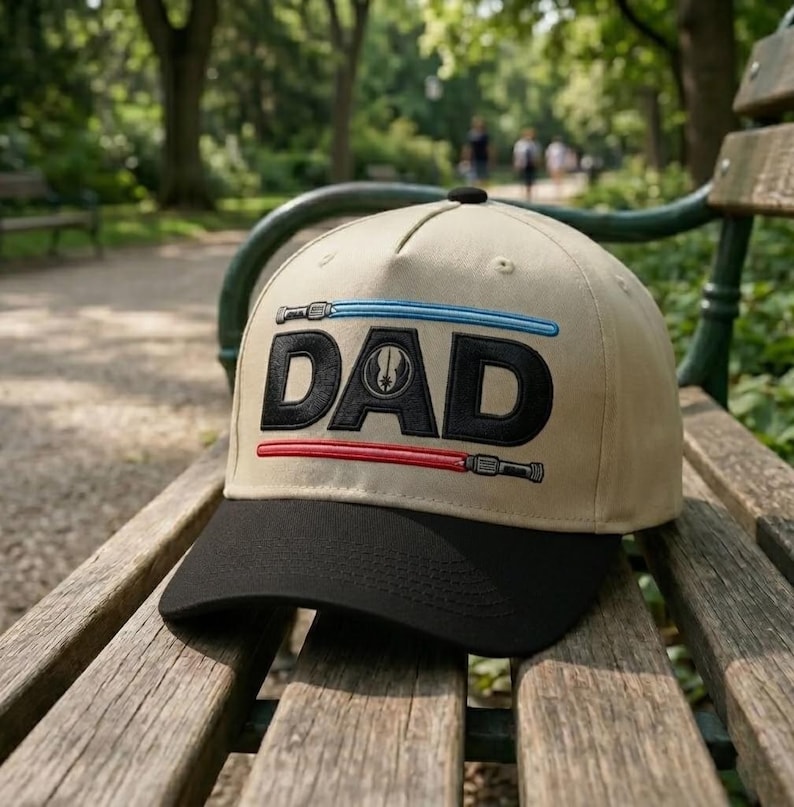 May include: A tan baseball cap with a black brim featuring the word "DAD" in black block letters. Above and below the word are embroidered lightsabers in blue and red. The cap is resting on a wooden bench in a park.