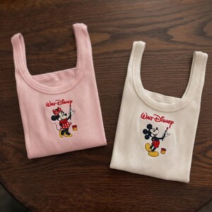 May include: Two ribbed tank tops, one pink and one white, featuring a Walt Disney logo and a cartoon character design. The pink tank top has a Minnie Mouse graphic, and the white tank top has a Mickey Mouse graphic.