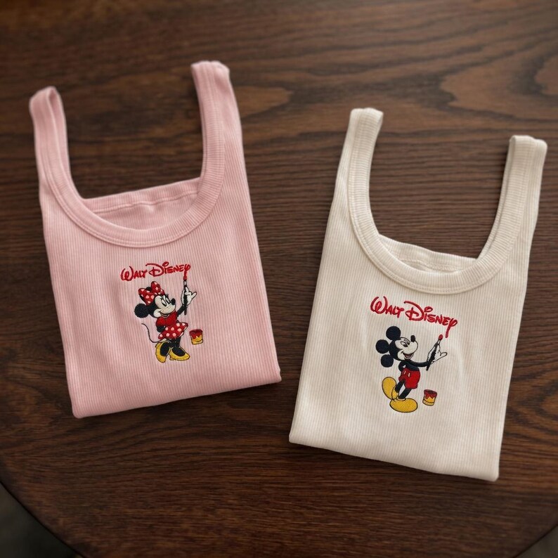 May include: Two ribbed tank tops, one pink and one white, featuring a Walt Disney logo and a cartoon character design. The pink tank top has a Minnie Mouse graphic, and the white tank top has a Mickey Mouse graphic.