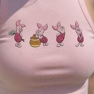 May include: Pink tank top featuring a Piglet design. The graphic shows Piglet in various poses, including holding flowers and a honey pot. The design is in a cartoon style.