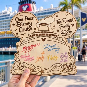 May include: A light-colored wooden plaque shaped like a Mickey Mouse head featuring the text Our First Disney Cruise and The Smith Family 2026. The center displays engraved signatures of Mickey Mouse, Donald Duck, Minnie Mouse, Goofy, Pluto, and Daisy Duck.