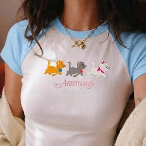 May include: A light blue and white raglan t-shirt featuring an embroidered graphic of three kittens from The Aristocats. The design includes an orange kitten, a gray kitten, and a white kitten with a pink bow, with the text The Aristocats below.