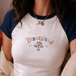 Puede incluir: Camiseta blanca de estilo b&eacute;isbol con mangas azul marino. La camiseta presenta la palabra "Disneyland" bordada en letras coloridas, junto con un gr&aacute;fico de Mickey Mouse y el a&ntilde;o "1955".