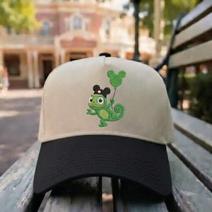 Puede incluir: Gorra de béisbol beige y negra con un camaleón verde bordado sosteniendo un globo verde de Mickey Mouse. El camaleón lleva un lazo negro y una diadema negra con orejas de Mickey Mouse. La gorra tiene una visera curva.