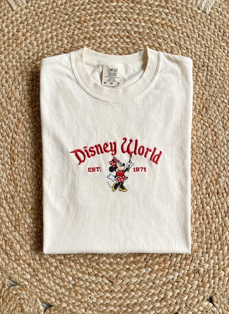 May include: Cream-colored t-shirt featuring the embroidered text "Disney World" in red script, with "EST. 1971" below. A Minnie Mouse graphic is in the center. The shirt is folded on a woven surface.