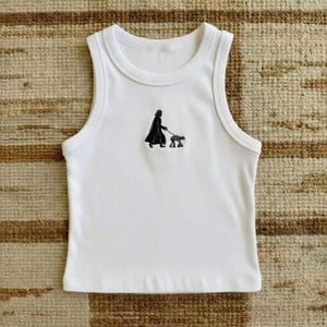May include: A white ribbed tank top featuring a small, centered black embroidered graphic of a cloaked figure walking a four-legged mechanical creature on a leash. The garment has a high, rounded neckline and a sleeveless, fitted silhouette.