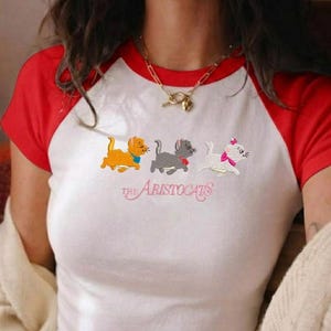 May include: A white raglan t-shirt with red sleeves featuring an embroidered graphic of three kittens from The Aristocats. The design includes an orange kitten, a gray kitten, and a white kitten with a pink bow, with the text The Aristocats below.