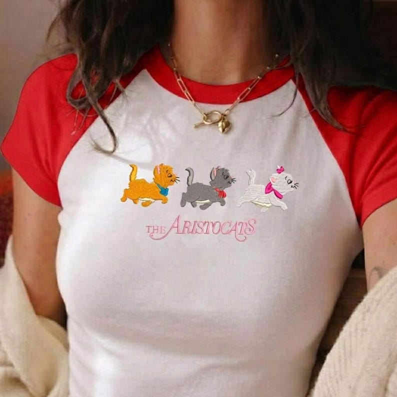 May include: A white raglan t-shirt with red sleeves featuring an embroidered graphic of three kittens from The Aristocats. The design includes an orange kitten, a gray kitten, and a white kitten with a pink bow, with the text The Aristocats below.