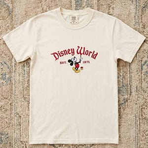 May include: Cream-colored t-shirt featuring the Disney World logo in red with Mickey Mouse. The graphic includes the text "EST. 1971". The shirt has short sleeves and a crew neck.