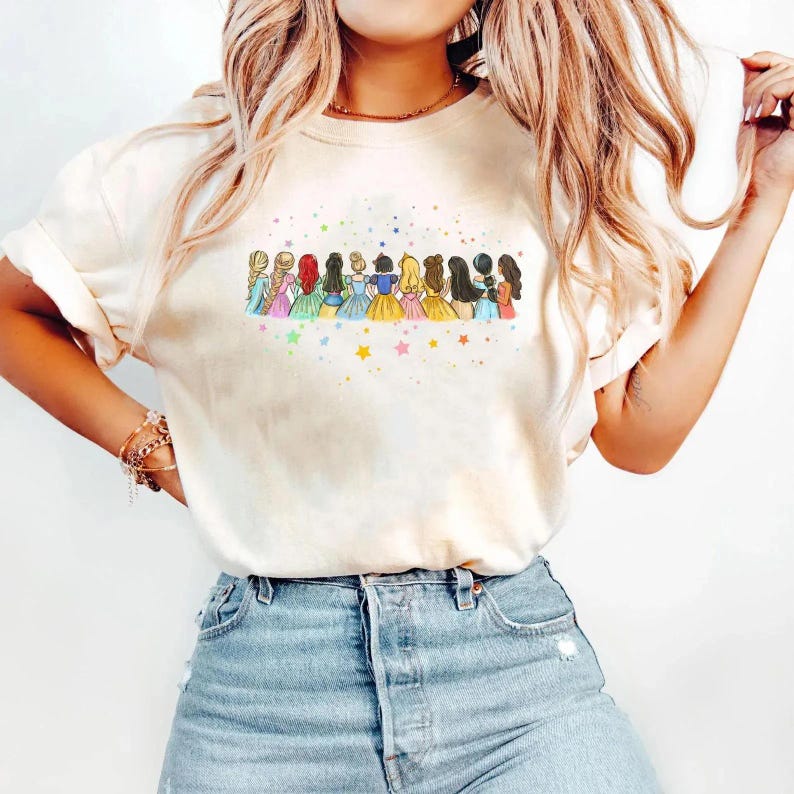 May include: A cream-coloured t-shirt with a colourful illustration of cartoon princesses. The princesses have different hairstyles and dresses, with stars above them. The t-shirt has rolled-up sleeves.