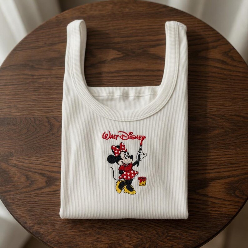 May include: White ribbed tank top featuring an embroidered Minnie Mouse design. The design includes Minnie Mouse holding a paintbrush, a paint can, and the words "Walt Disney" in red script. The tank top is folded neatly.
