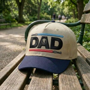 May include: A tan baseball cap with a navy blue brim. The word "DAD" is embroidered in black, with blue and red lightsaber designs above and below the text. The cap is resting on a wooden bench in a park.