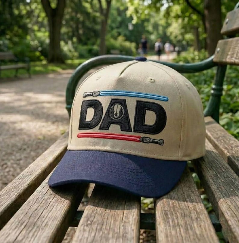 May include: A tan baseball cap with a navy blue brim. The word "DAD" is embroidered in black, with blue and red lightsaber designs above and below the text. The cap is resting on a wooden bench in a park.