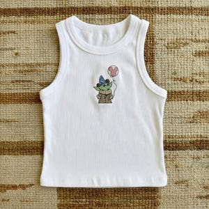 Puede incluir: Camiseta sin mangas blanca con un dibujo animado de un personaje verde con un sombrero azul y una capa marr&oacute;n, sosteniendo un globo rosa de Mickey Mouse. La camiseta sin mangas es sin mangas y tiene un cuello redondo.