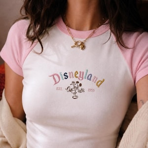 Puede incluir: Camiseta ragl&aacute;n blanca y rosa con texto bordado "Disneyland" y gr&aacute;fico de Mickey Mouse. El texto es multicolor, con "EST. 1955" debajo. Tambi&eacute;n se ve un collar dorado.