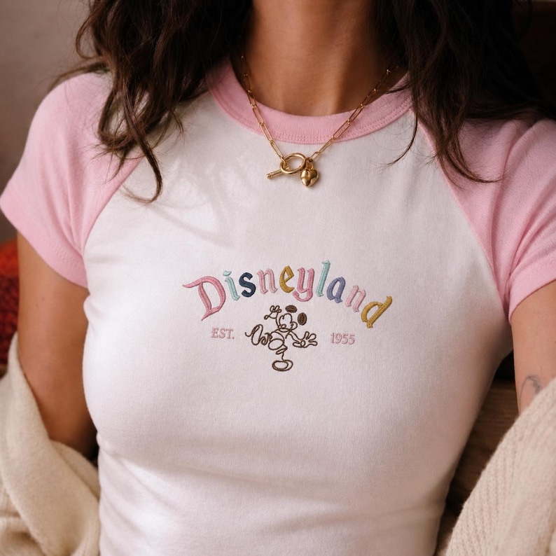 Puede incluir: Camiseta ragl&aacute;n blanca y rosa con texto bordado "Disneyland" y gr&aacute;fico de Mickey Mouse. El texto es multicolor, con "EST. 1955" debajo. Tambi&eacute;n se ve un collar dorado.