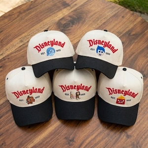 Puede incluir: Se muestran cinco gorras de béisbol blancas con viseras negras. Cada gorra presenta la palabra "Disneyland" en rojo, el año "1955" y un personaje de dibujos animados. Están dispuestas sobre una superficie de madera.