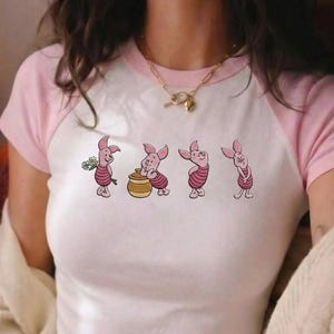 May include: White and pink raglan t-shirt featuring a cartoon illustration of Piglet from Winnie the Pooh. The design shows Piglet in four different poses. The shirt has a pink neckline and sleeves.
