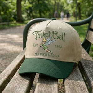 Puede incluir: Una gorra de béisbol beige con visera verde oscuro que presenta un gráfico bordado de Tinker Bell. El texto Tinker Bell, Est. 1953 y Neverland está bordado en hilo verde. La gorra tiene una corona estructurada y detalles de costura.