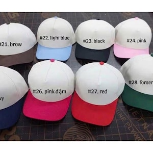 May include: A collection of baseball caps with white crowns and colored brims. The caps are labeled with numbers and colors, including brown, light blue, black, pink, navy, pink, red, and forest green.