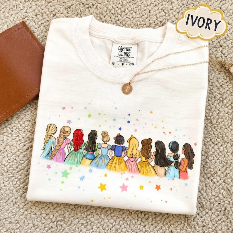 May include: Ivory t-shirt with a cartoon illustration of princesses in colourful dresses and hairstyles. A gold necklace with a pendant lies on the shirt. The shirt has a Comfort Colors label.