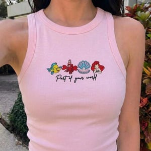 May include: Pink tank top with embroidered cartoon characters and the text "Part of your world". The design features a yellow and blue fish, a red crab, a blue and white seashell, and a red-haired mermaid.