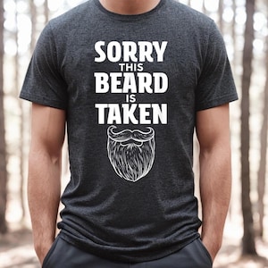 Peut inclure: Un t-shirt gris foncé avec le texte "Désolé, cette barbe est prise" en lettres blanches et une barbe de dessin animé blanche avec une moustache en dessous du texte.
